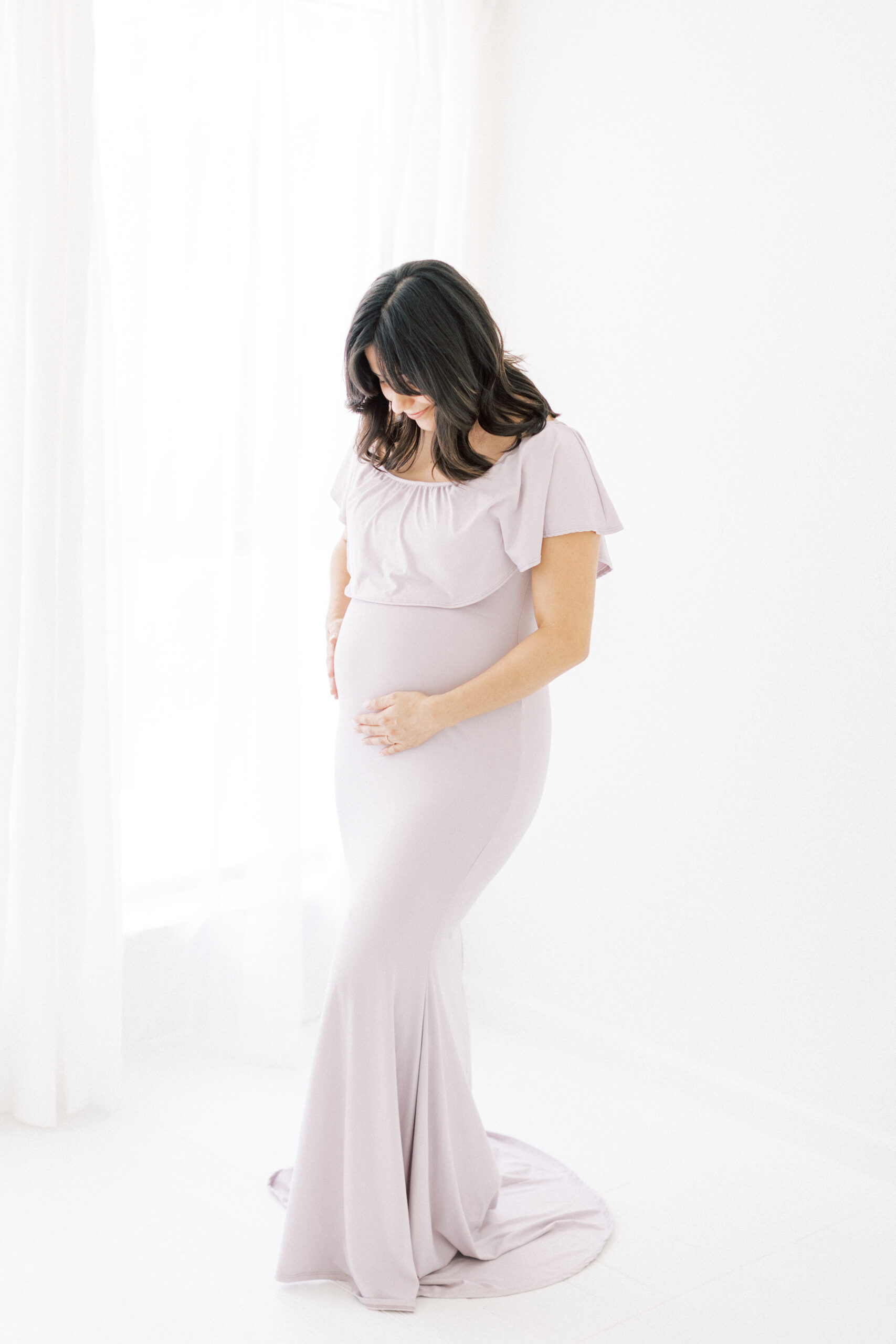A mom to be stands in a purple dress looking down at her bump in a studio by a window