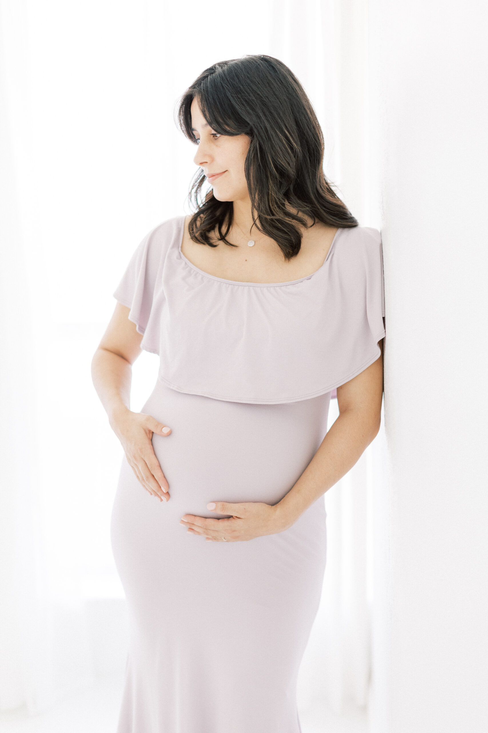 A mother to be in a maternity gown leans against a wall in front of a window in a studio Dallas Midwife Associates