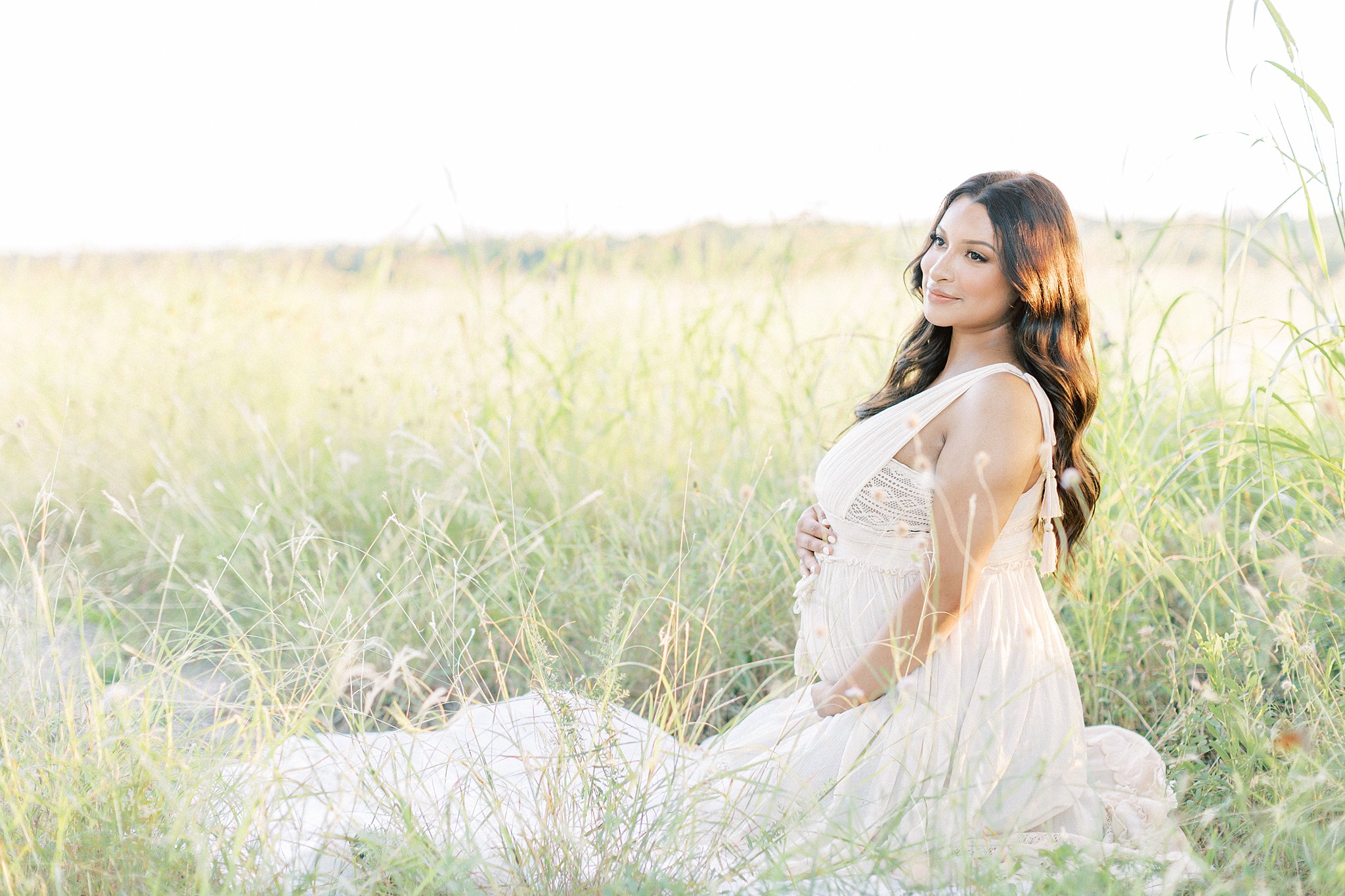 A mother to be kneels in a field of tall grass in a white maternity dress Dallas Prenatal Yoga
