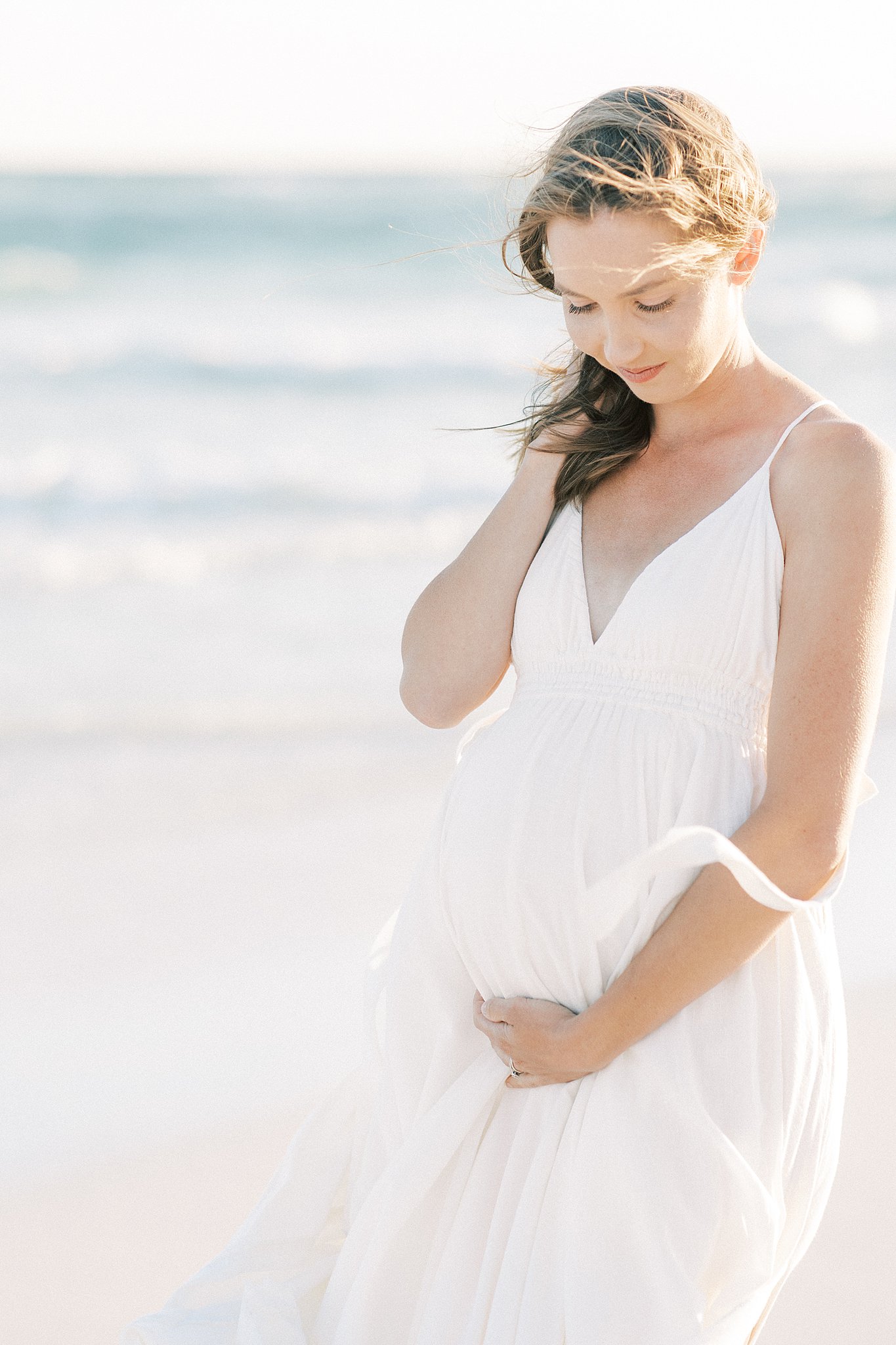 A mom to be stands on a windy beach in a white maternity dress holding her bump Childbirth Classes Dallas