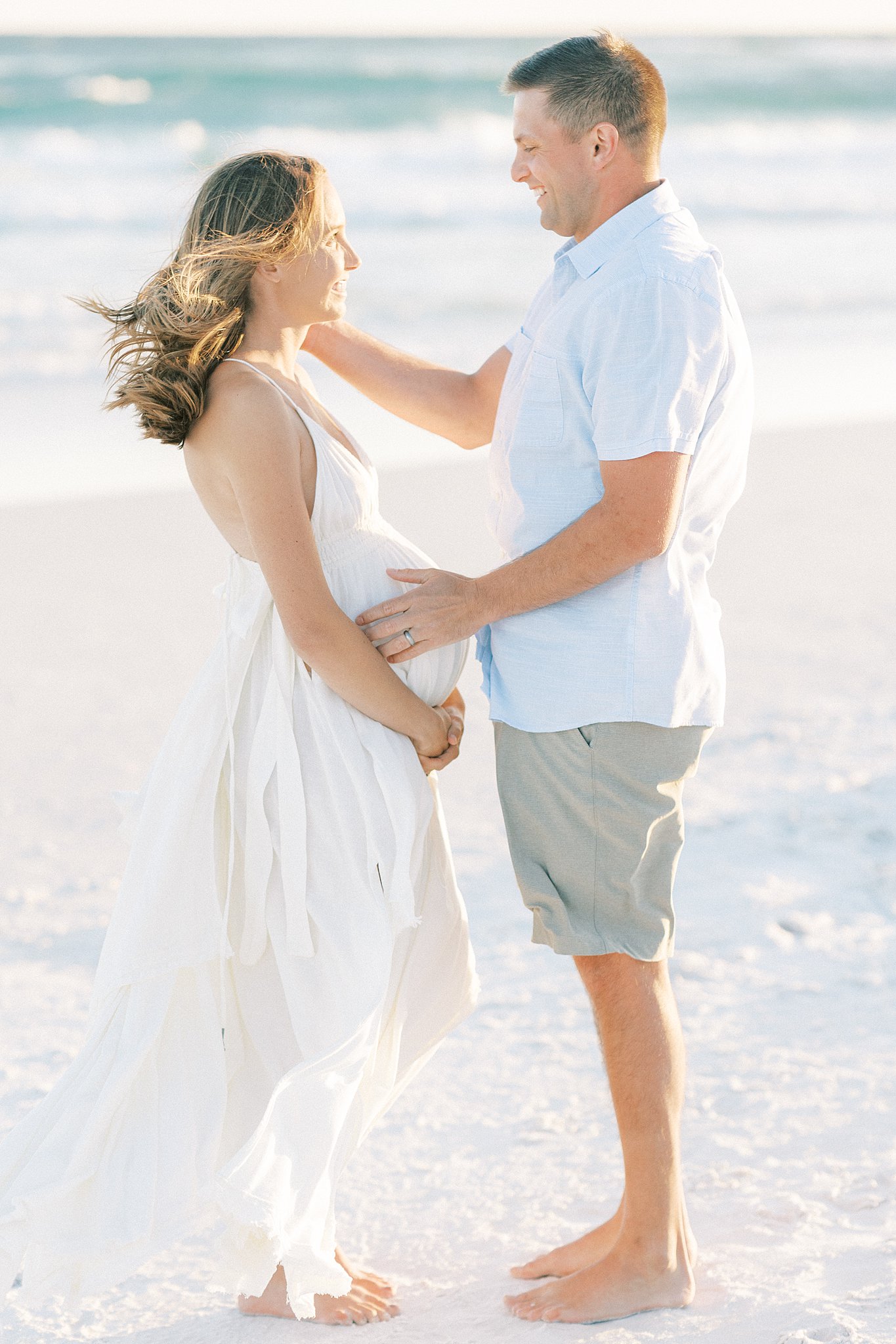 A mom to be in a white maternity dress stands on the beach with her husband running a hand through her hair Childbirth Classes Dallas