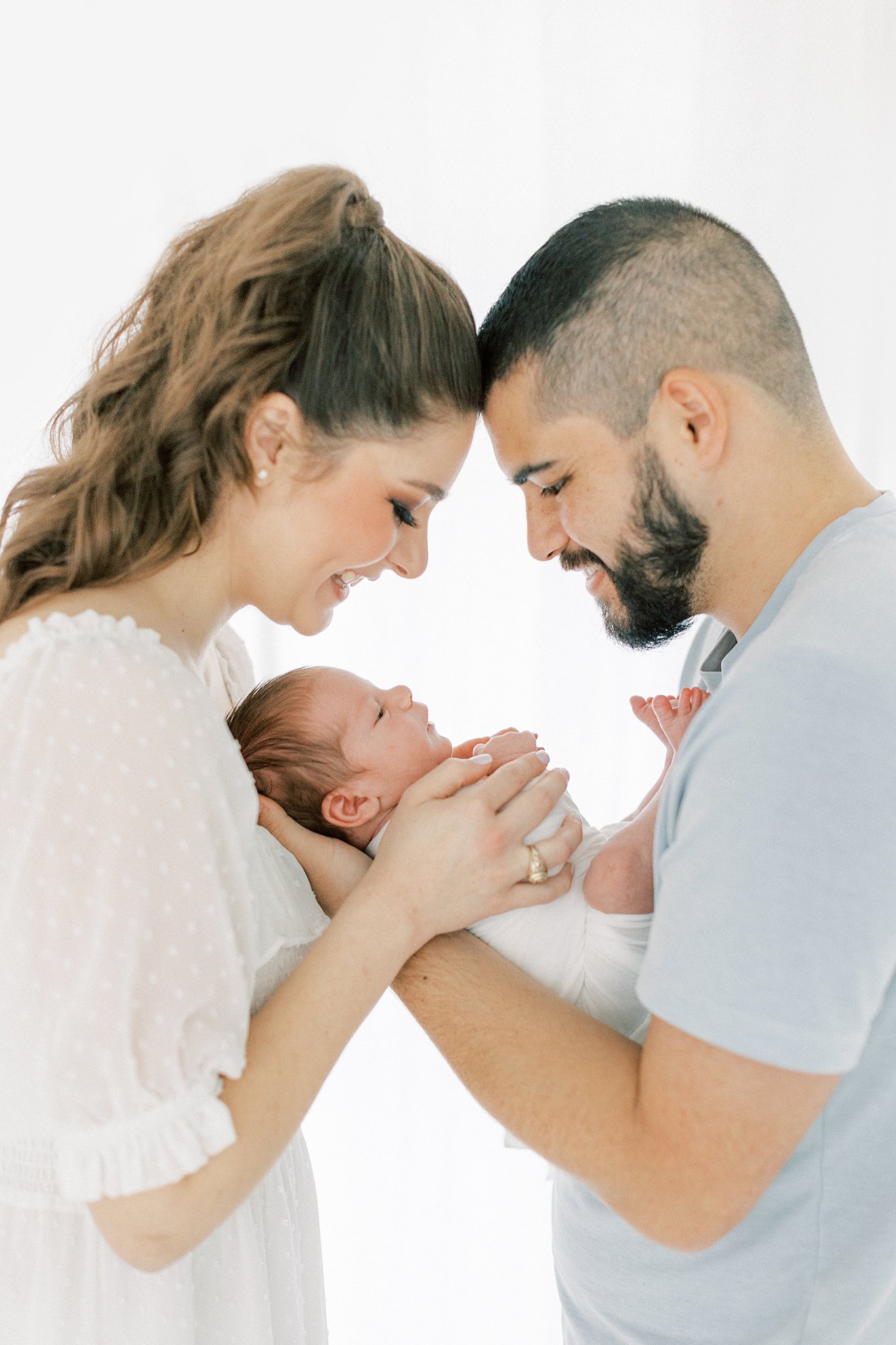New parents smile down at their newborn baby while standing with foreheads together in front of a window