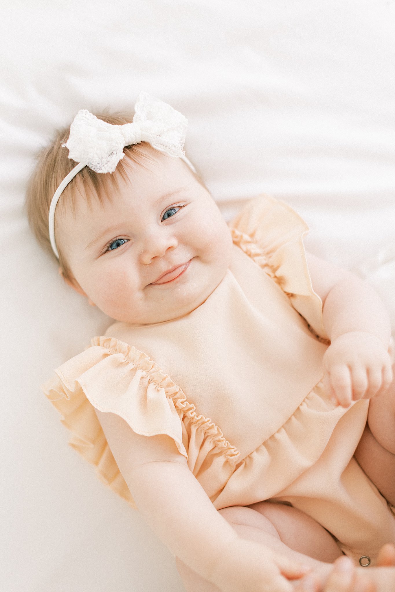 An infant baby in a peach dress lays on a bed smiling and sticking her tongue out