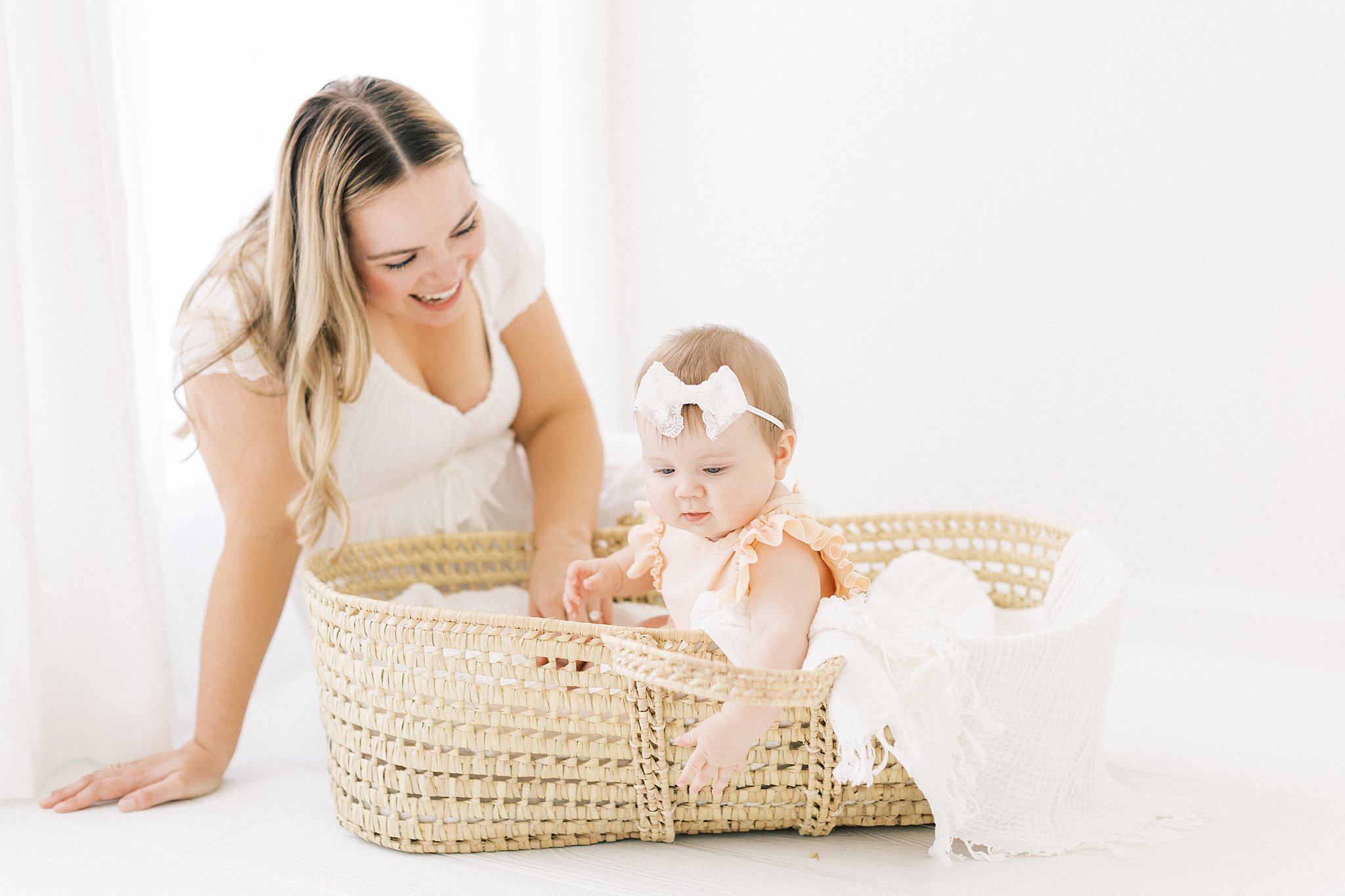 An infant baby girl sits and plays in a woven basket with a lace bow on her head while mom sits behind her Dondolo Dallas