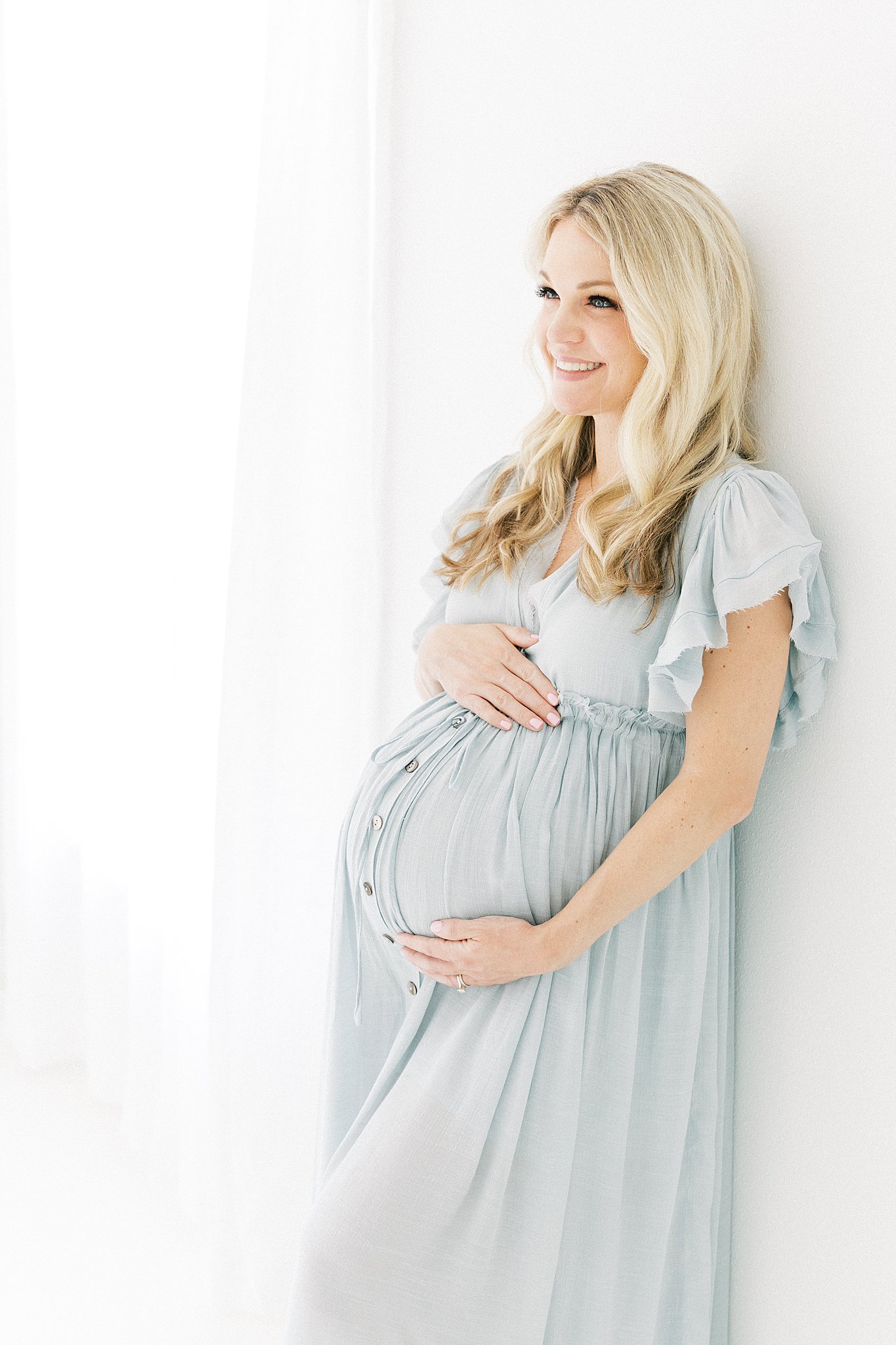 A mom to be leans against a wall smiling in a blue maternity dress holding her bump in a studio