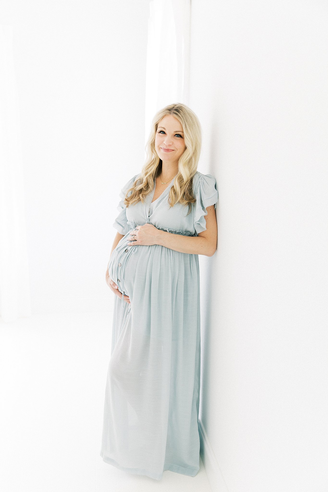 A mom to be leans against a wall in a white studio by a window holding her bump in a blue maternity dress after visiting Lovers Lane Birth Center