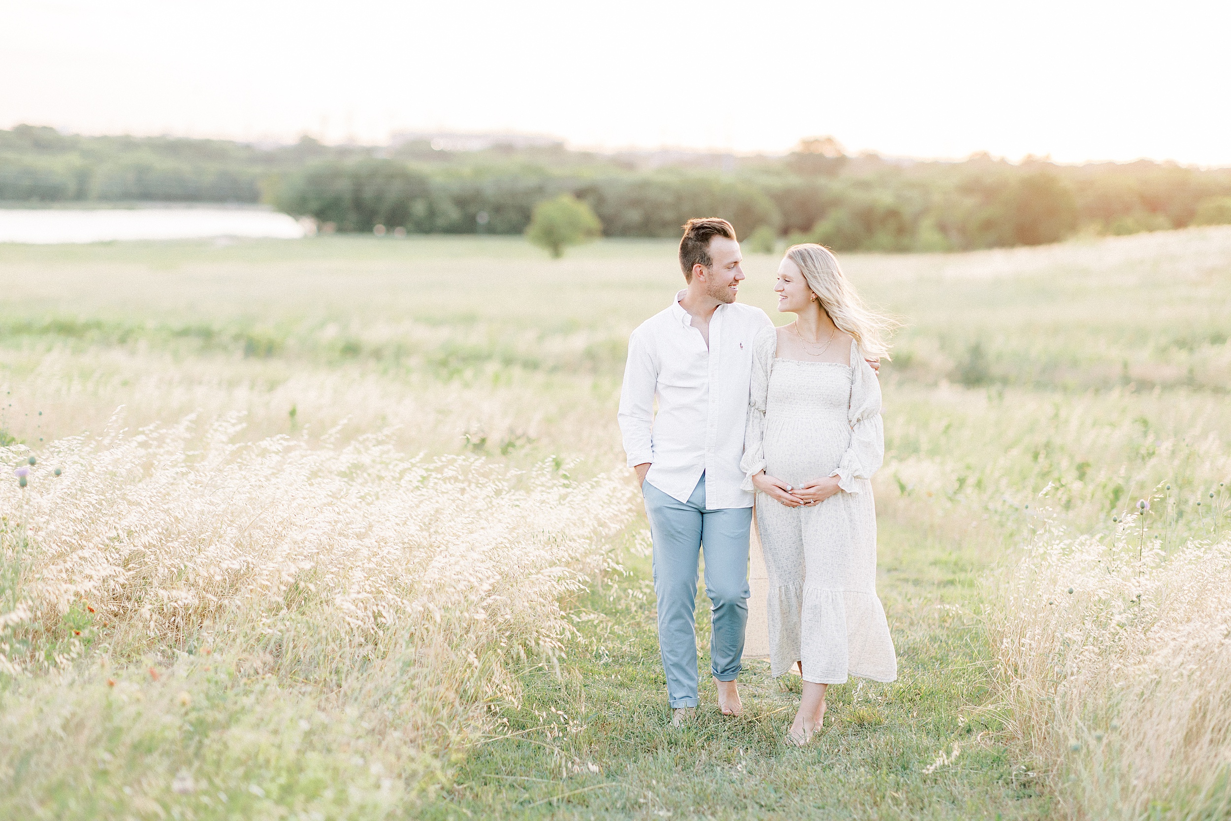A mom to be holds her bump while walking through a field of tall grass with her partner in blue pants and a white shirt