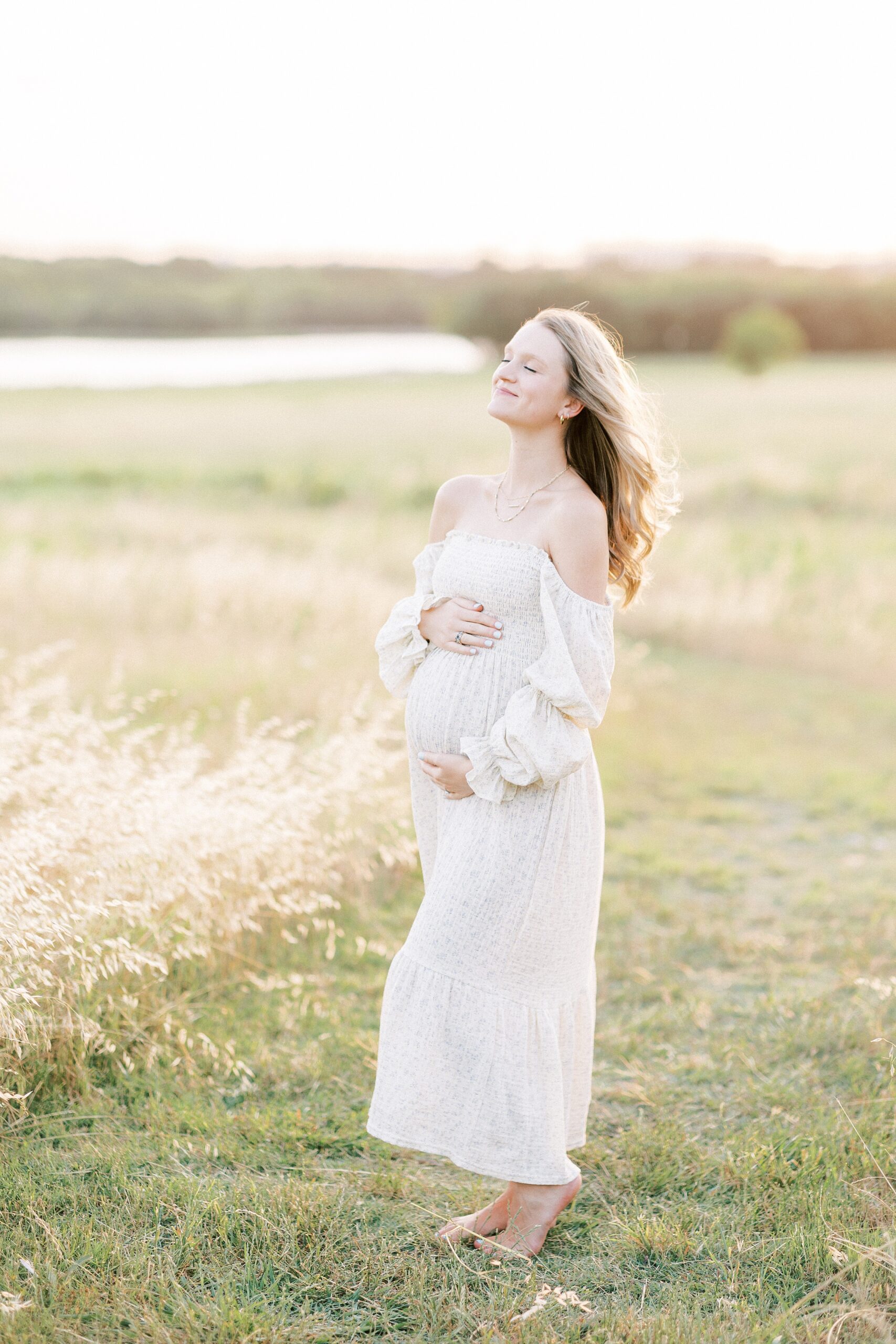 A mom to be basks in the sun and wind in a field of tall grass by a lake in a yellow maternity dress with hands on her bump Origins Birth And Wellness