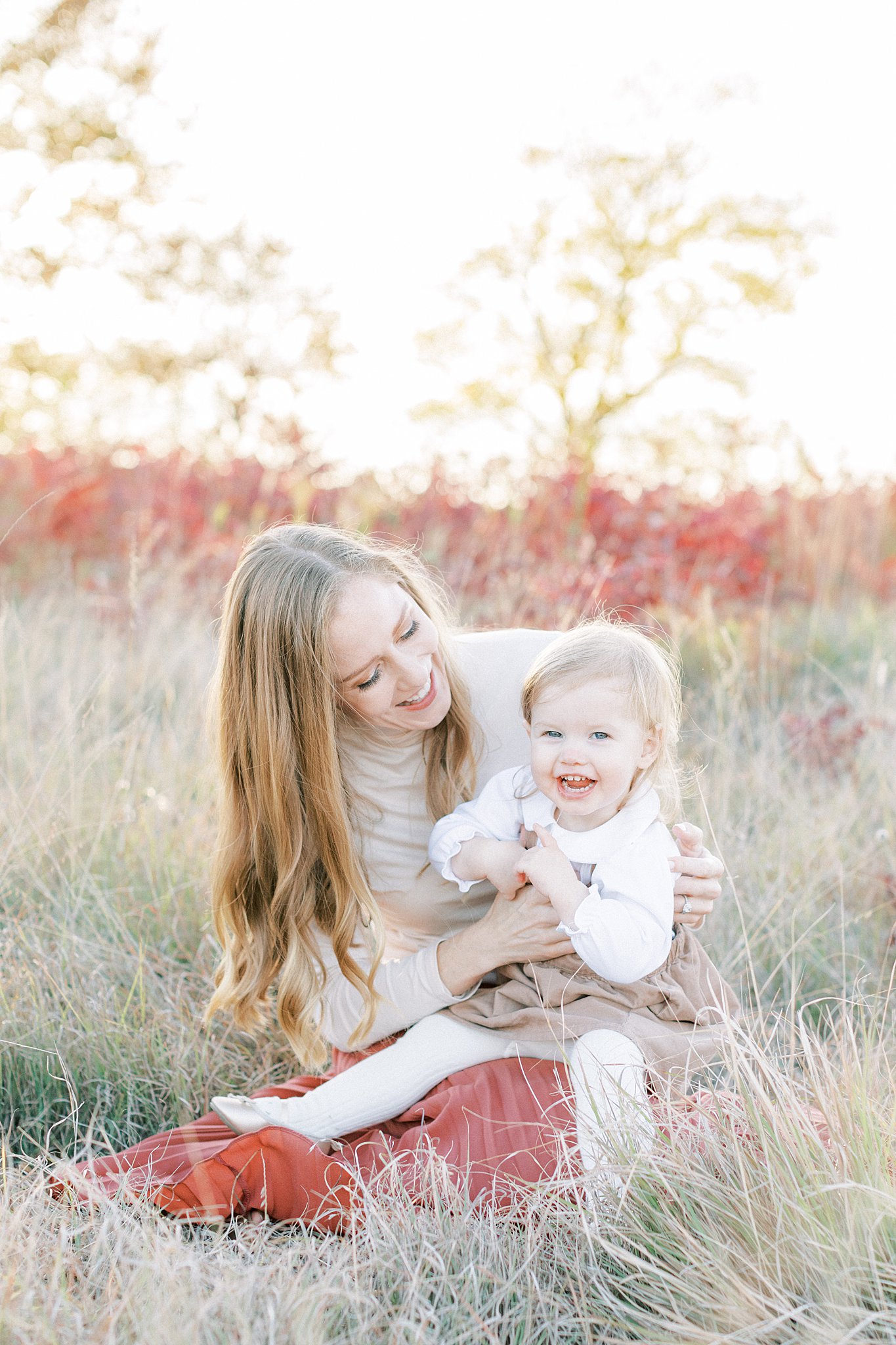 A young girl in a brown dress smiles and plays while sitting in mom's lap in a field of tall grass at sunset Things to do in Dallas with Toddlers