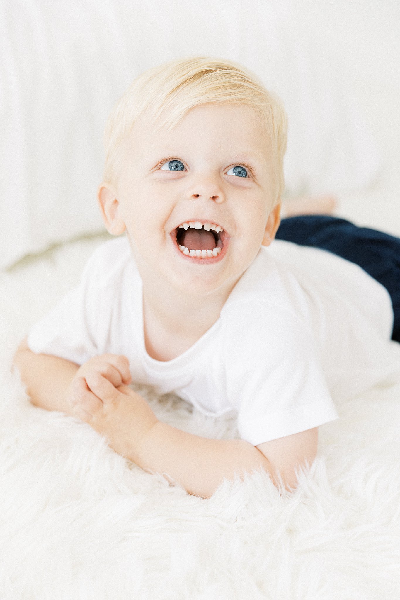 A toddler boy lays on a white rug smiling wide and wearing a white shirt