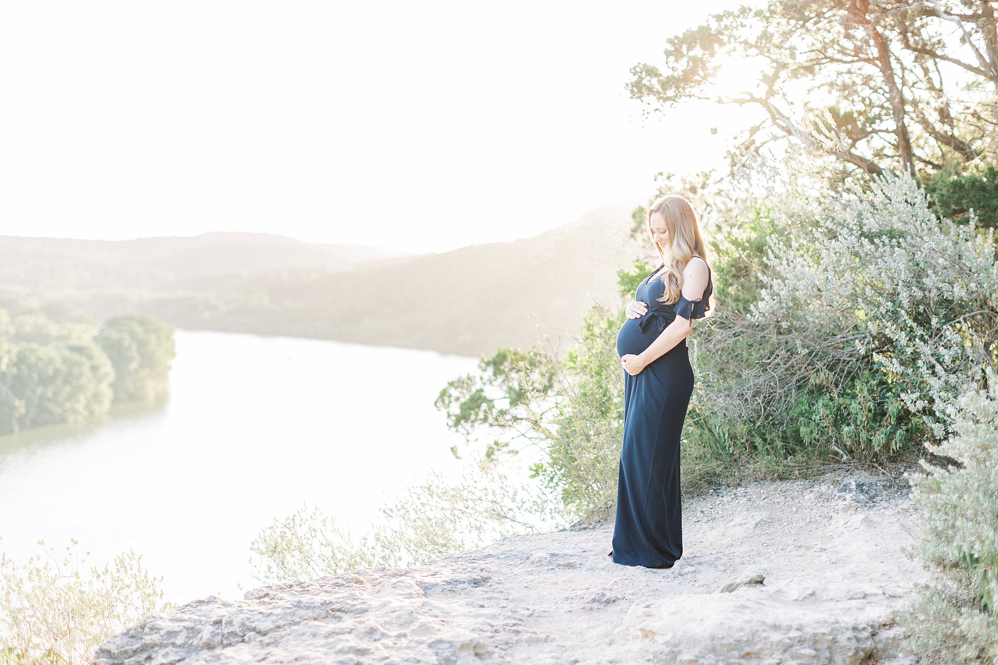 A mom to be stands on a mountain path overlooking a river and holding her bump in a blue maternity dress after visiting Bundleborn Midwifery