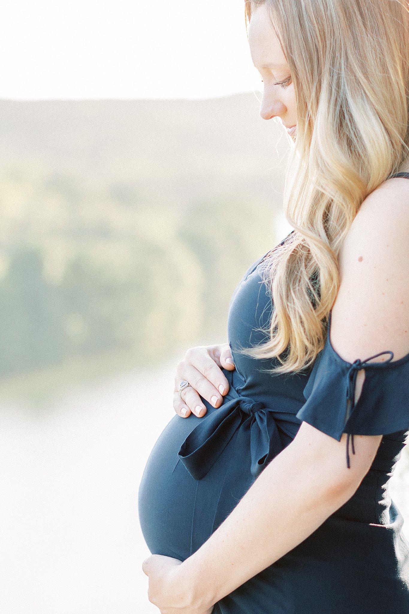 A mom to be stands in the sun holding her bump in a blue maternity dress after visiting Bundleborn Midwifery