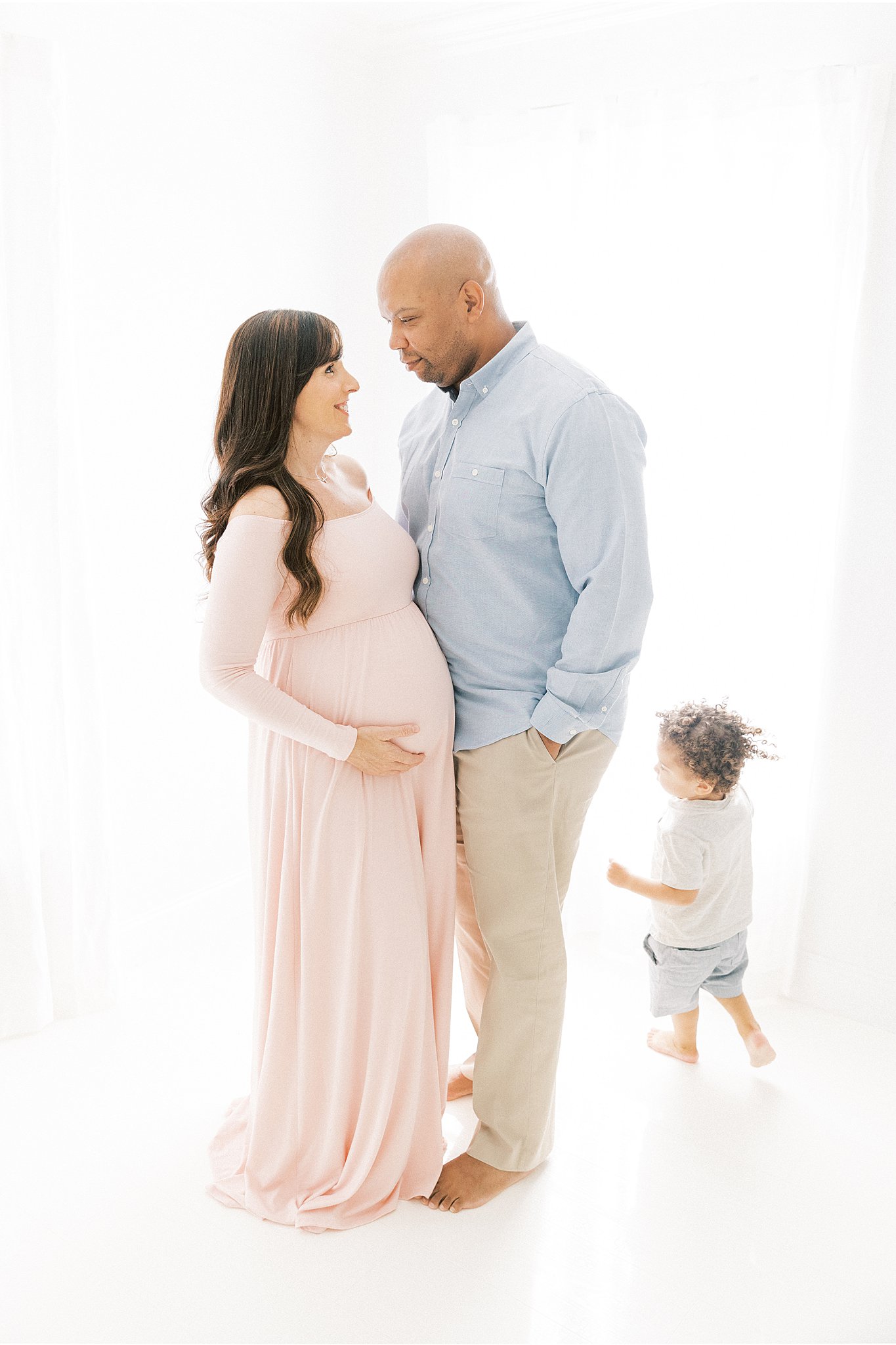 A mom to be stands in a pink maternity gown with her husband while their toddler runs around them after visiting Frisco Birth Center