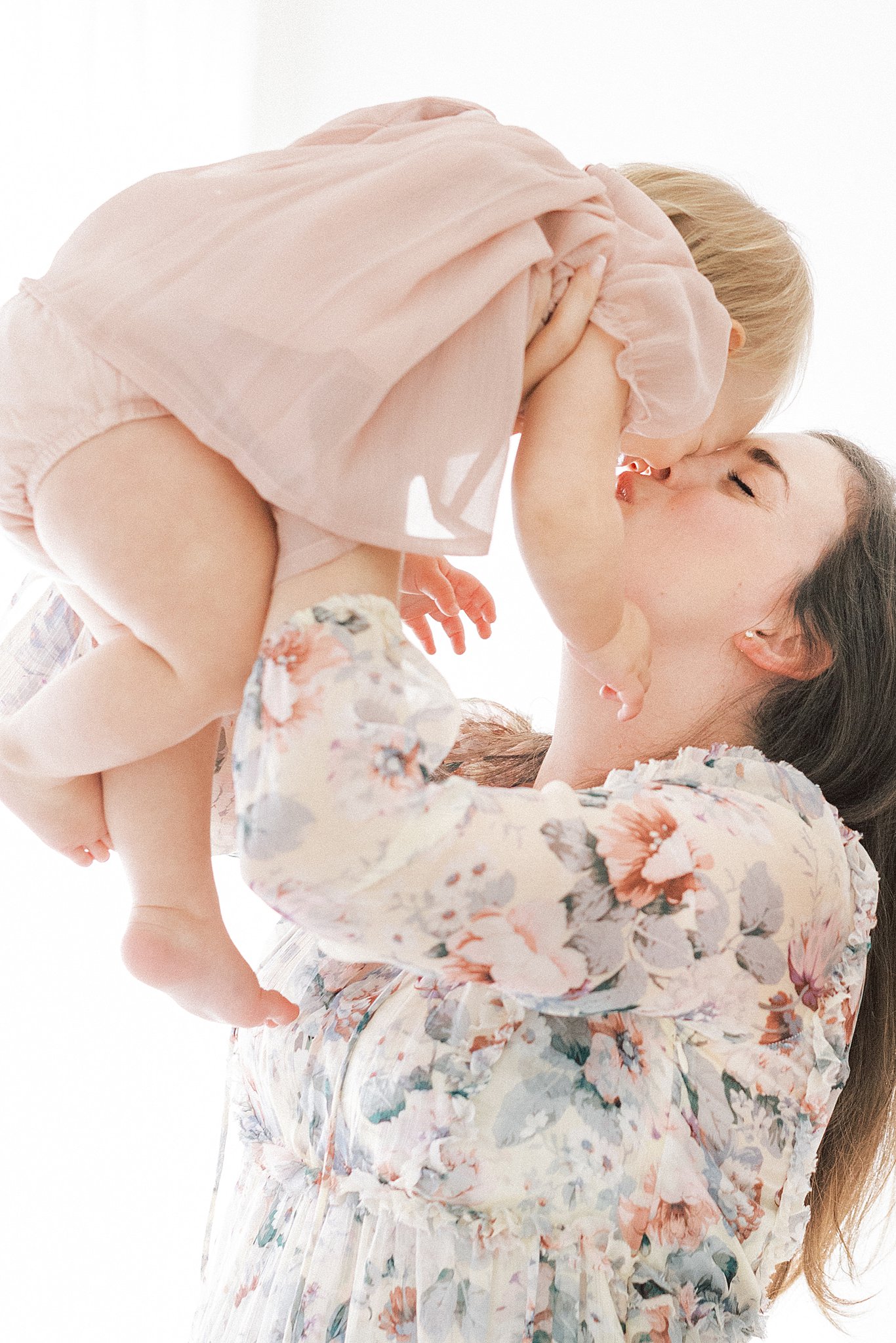 A mother in a floral dress lifts and kisses her toddler daughter in a studio