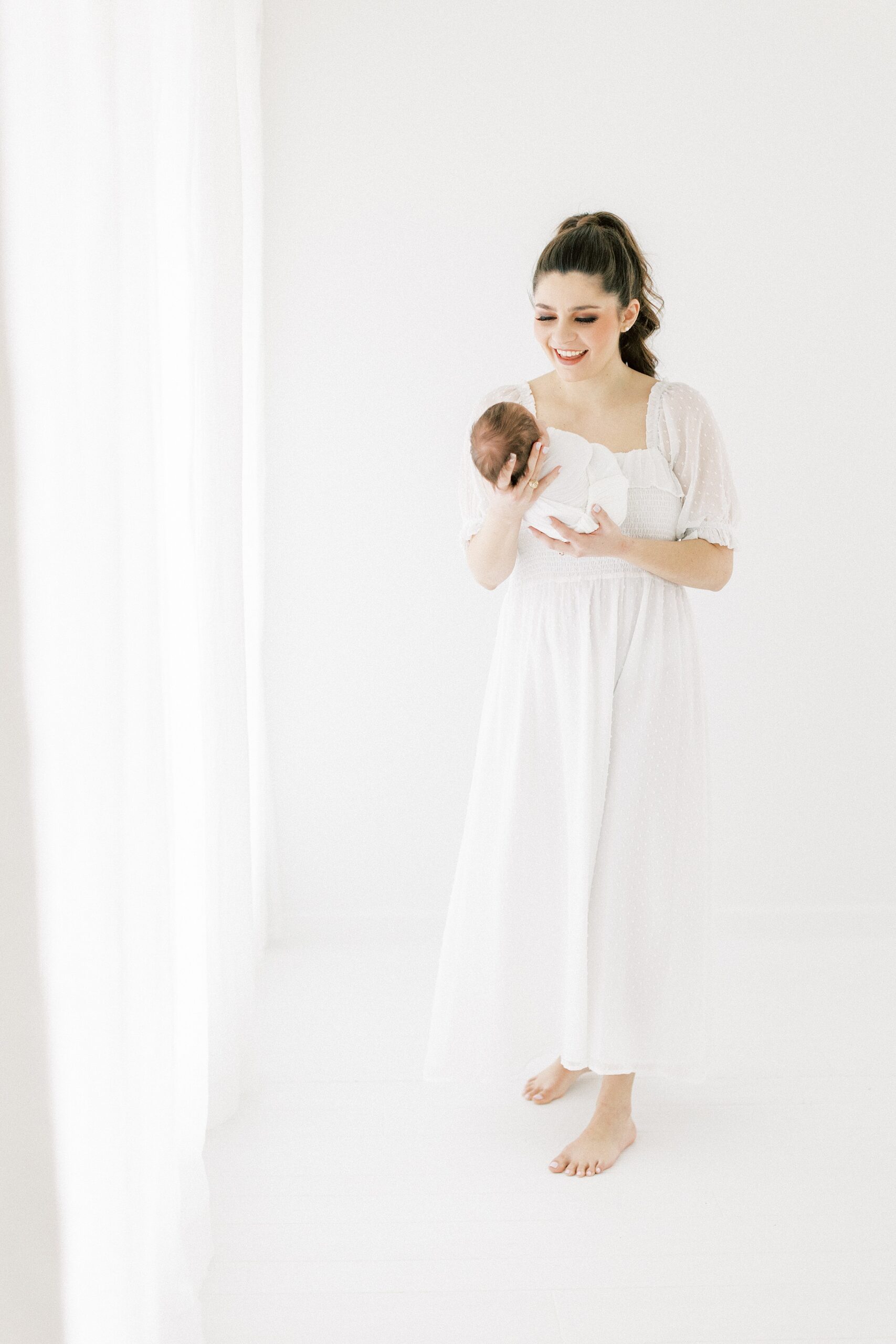A happy mother in a white dress stands in a studio holding her newborn baby in front of her after meeting a Dallas Au Pair