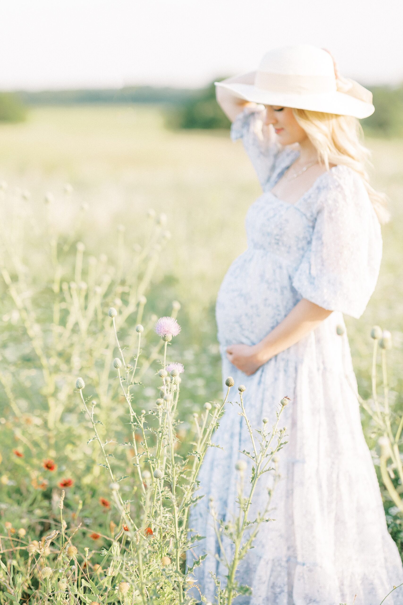 A mother to be walks through a field of wildflowers holding her bump and hat in a blue floral dress after using Dallas Fertility Clinics