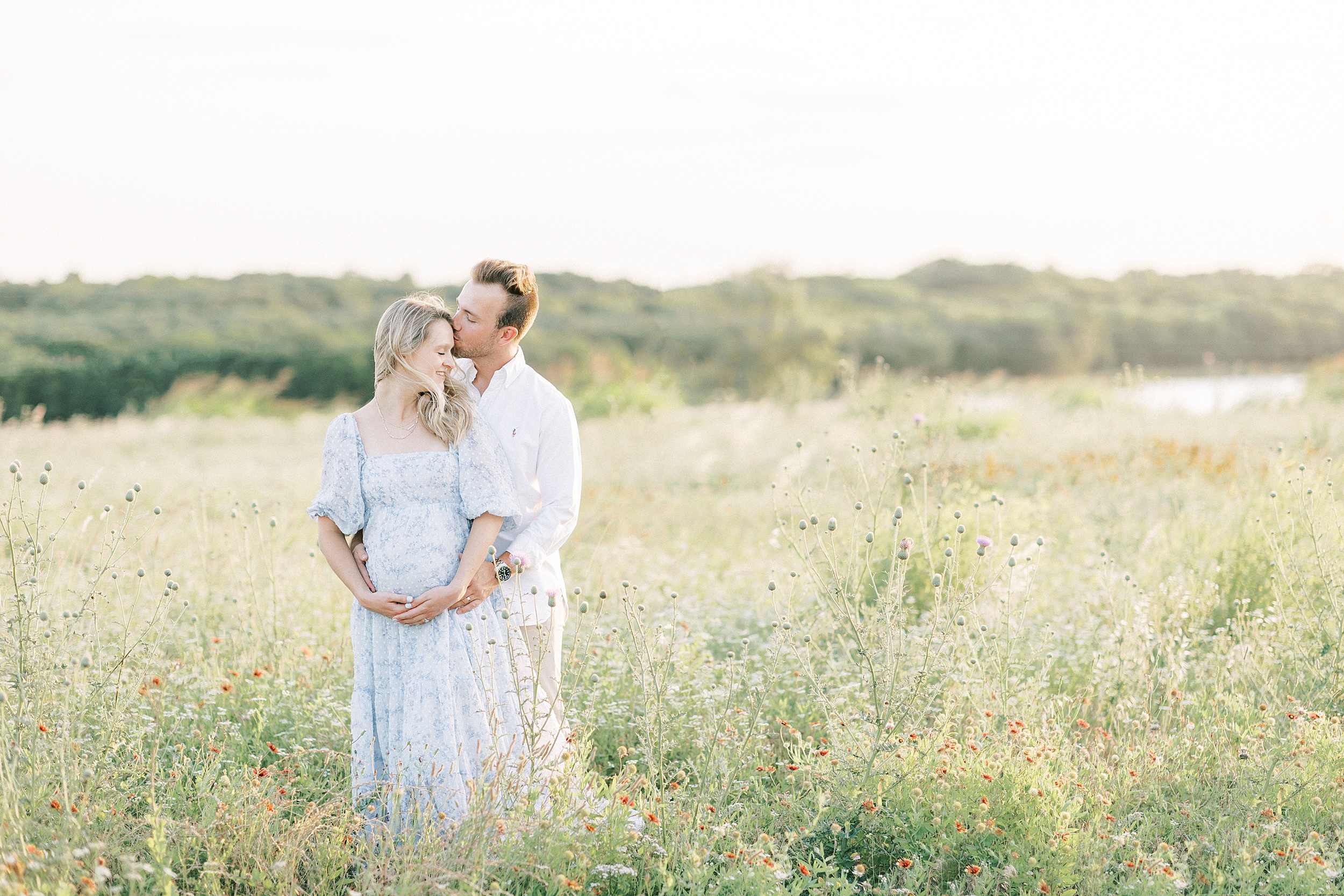 A mother to be in a blue floral dress stands in a field of wildflowers at sunset while being hugged and kissed by her husband after using Dallas Fertility Clinics