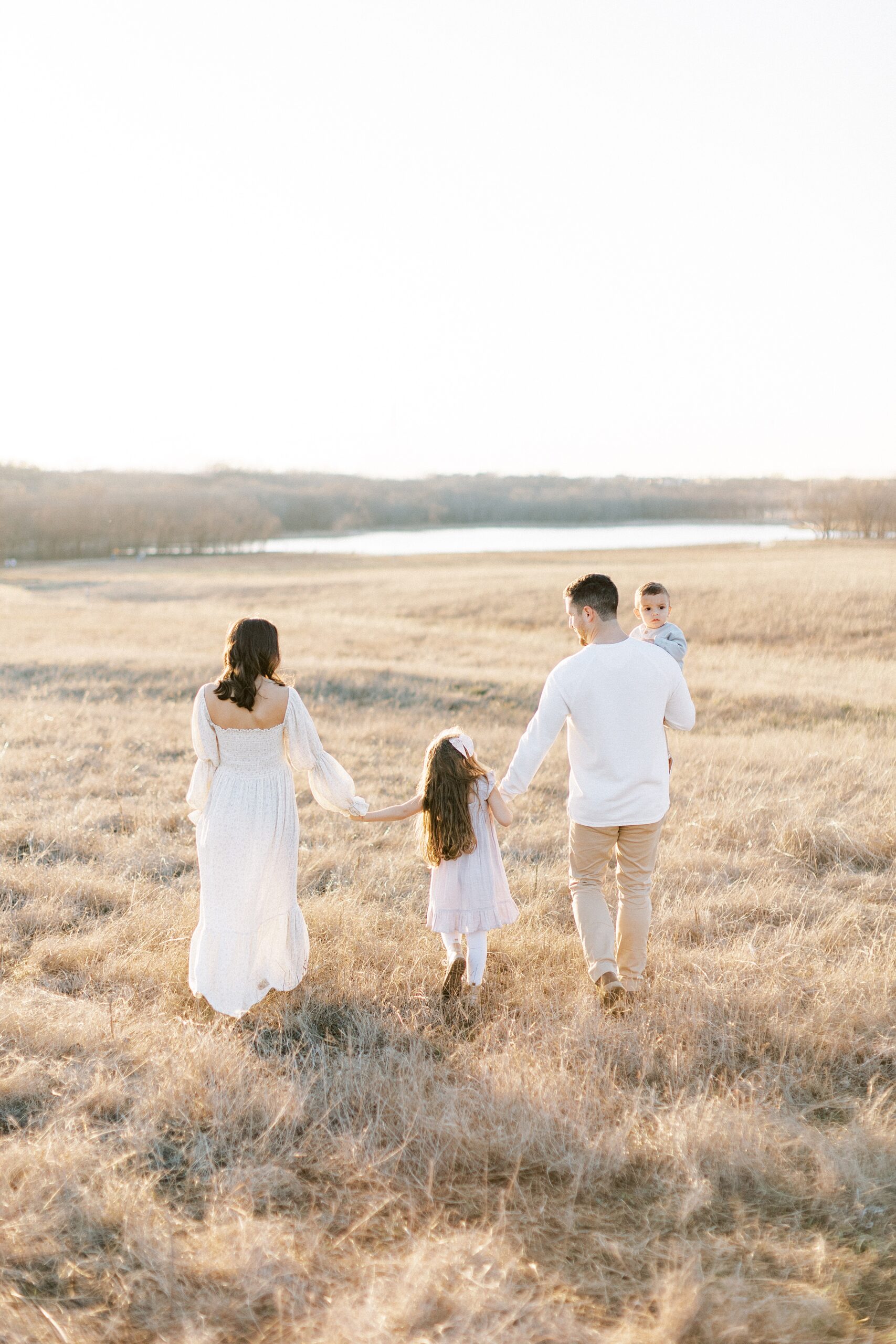 A mom and dad walk through a large field to a lake holding hands with their toddler daughter and holding their toddler son after visiting Dallas Pediatricians