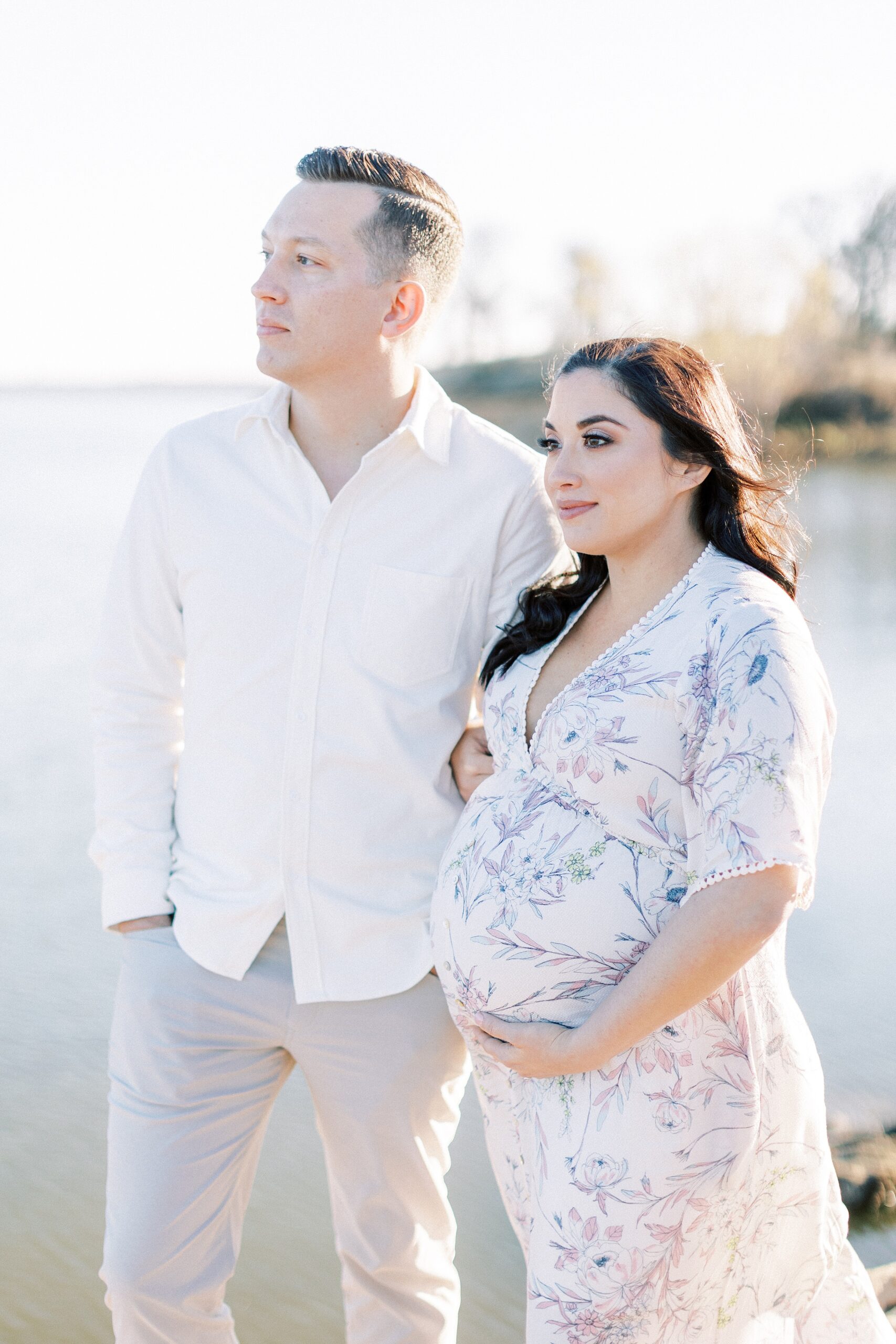 A mother to be in a floral printed maternity dress stands on a beach looking out to the water with her husband after meeting a Postpartum Doula Dallas