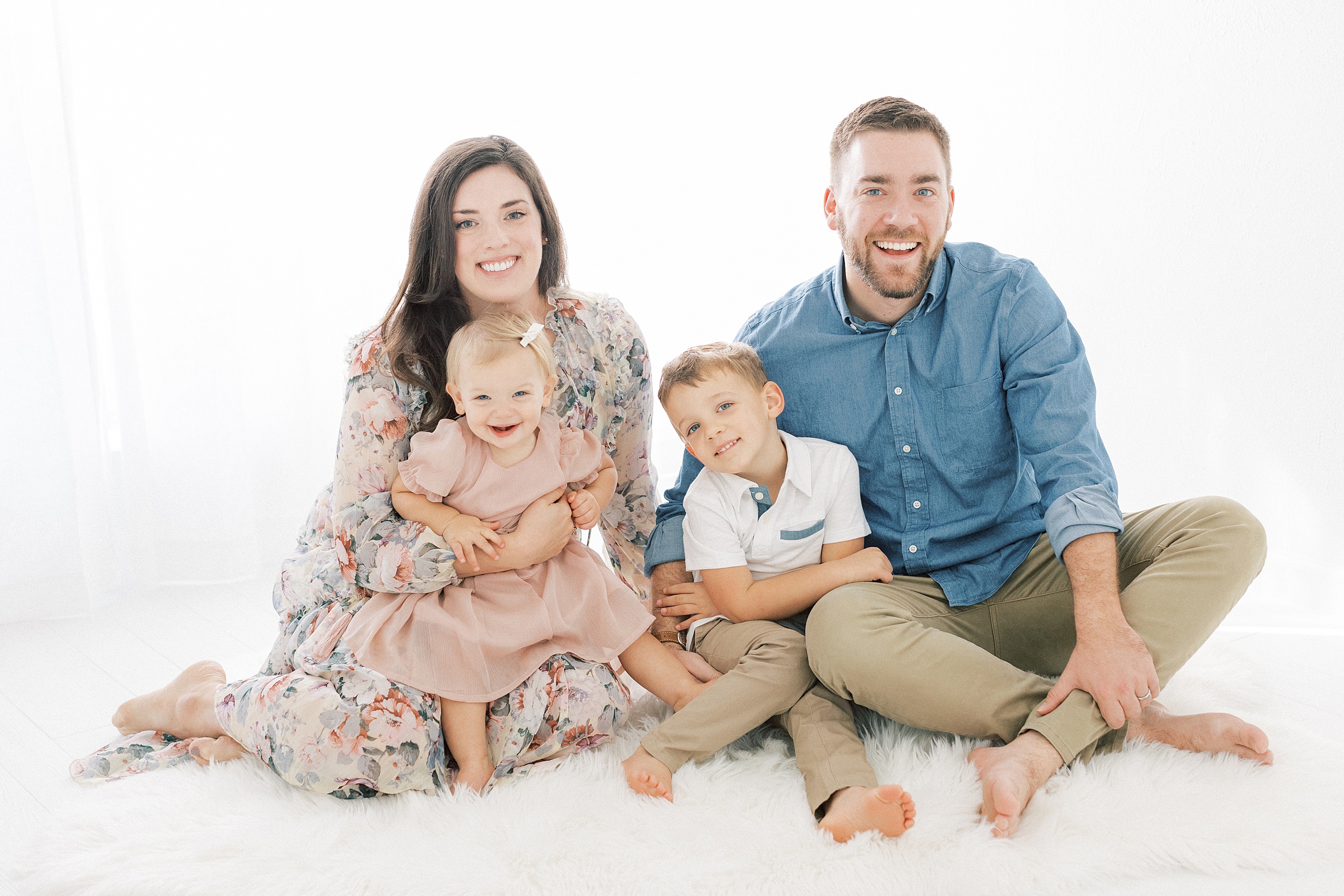 A mom and dad sit on the floor of a studio with their toddler son and daughter in their laps