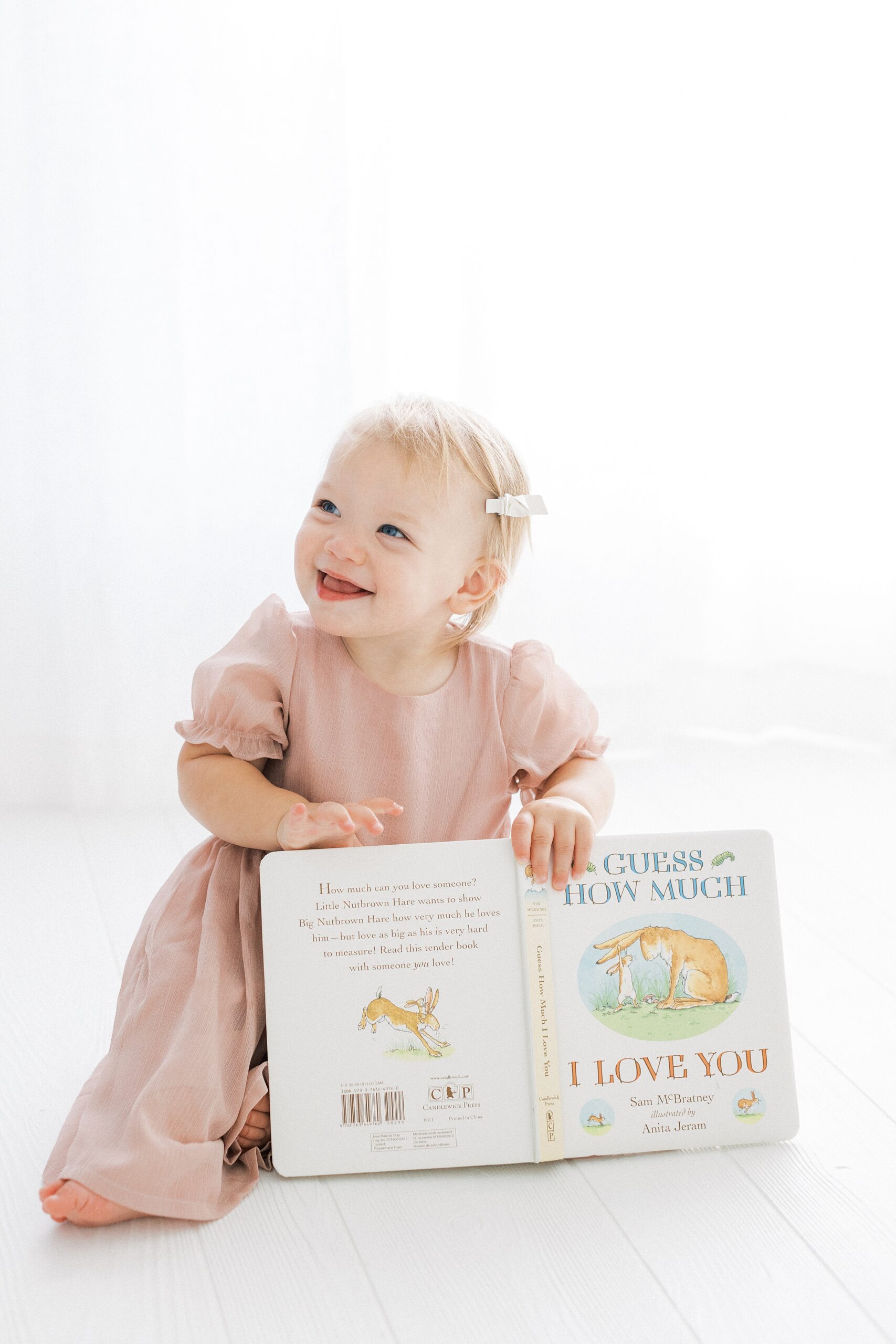 A happy young toddler girl in a pink dress sits on the floor of a studio holding a book from Cultivated Kids