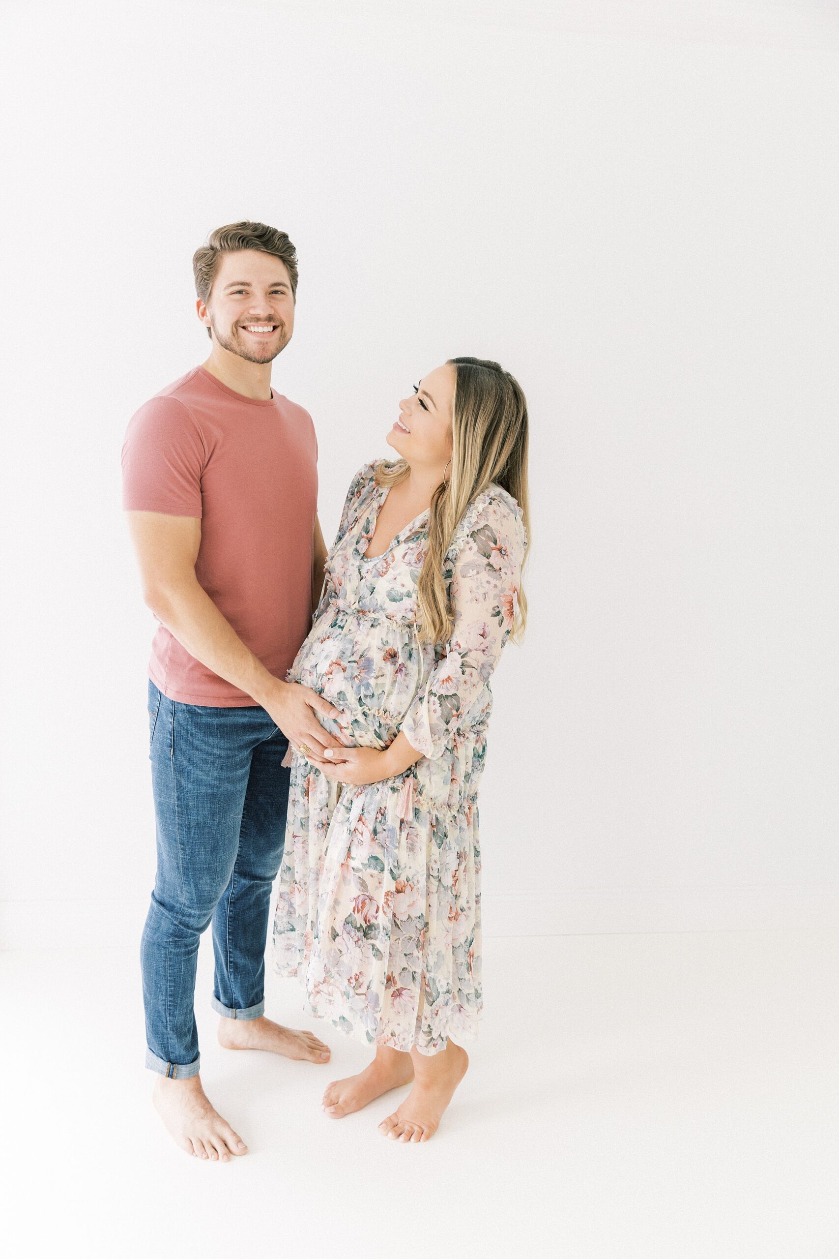 A mother to be in a floral print dress holds her bump with her husband while standing in a studio