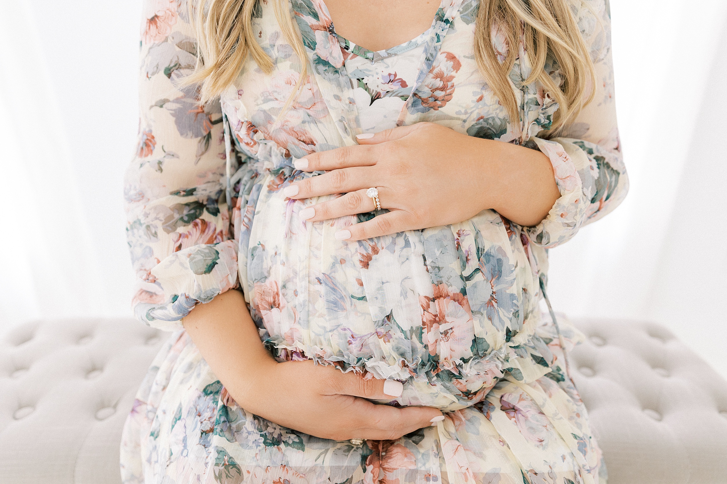 A mother to be in a floral maternity dress holds her bump while sitting on a bench in a studio before visiting Plano Birth Center