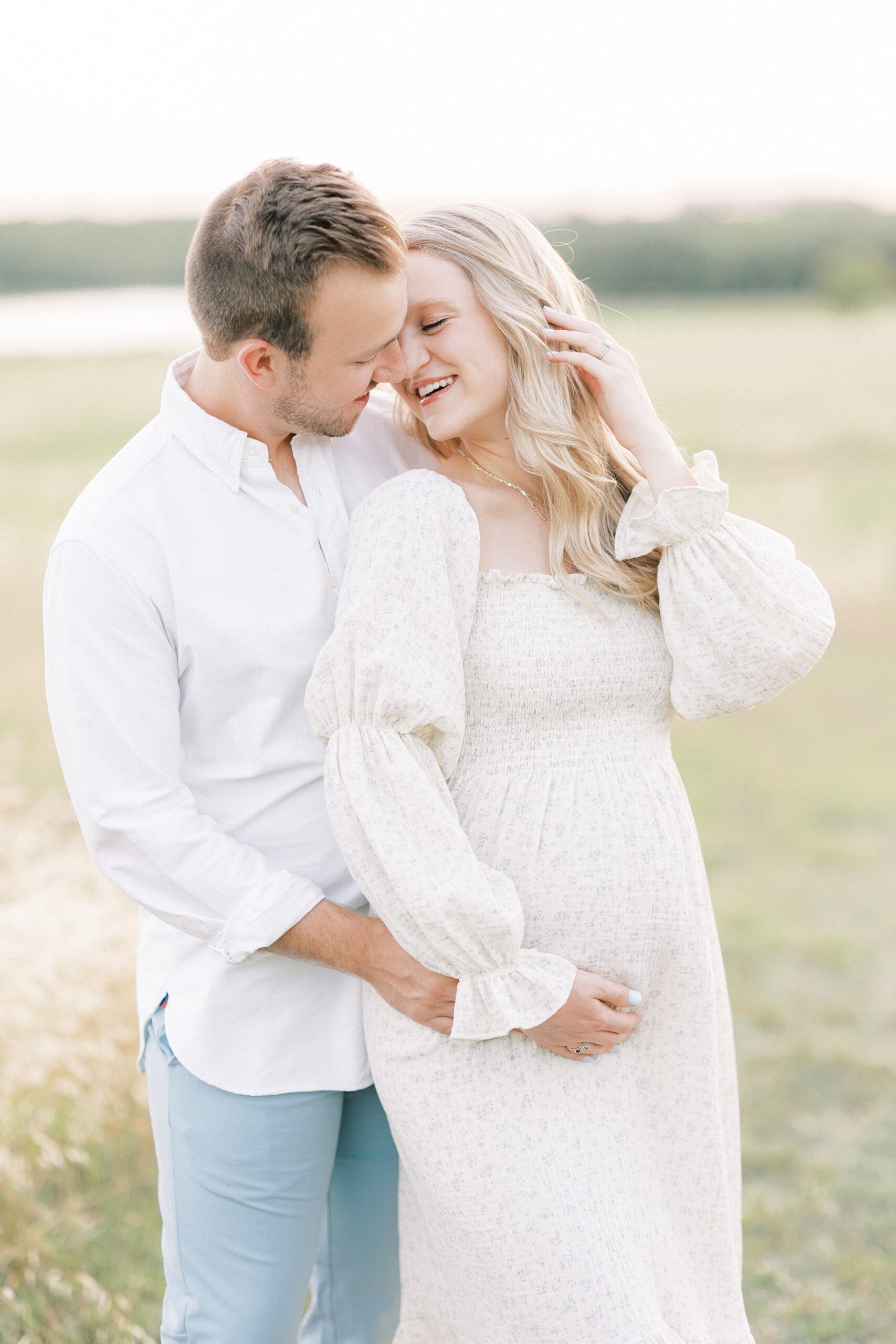 Expecting parents hold the bump while leaning in for a kiss in a windy field at sunset