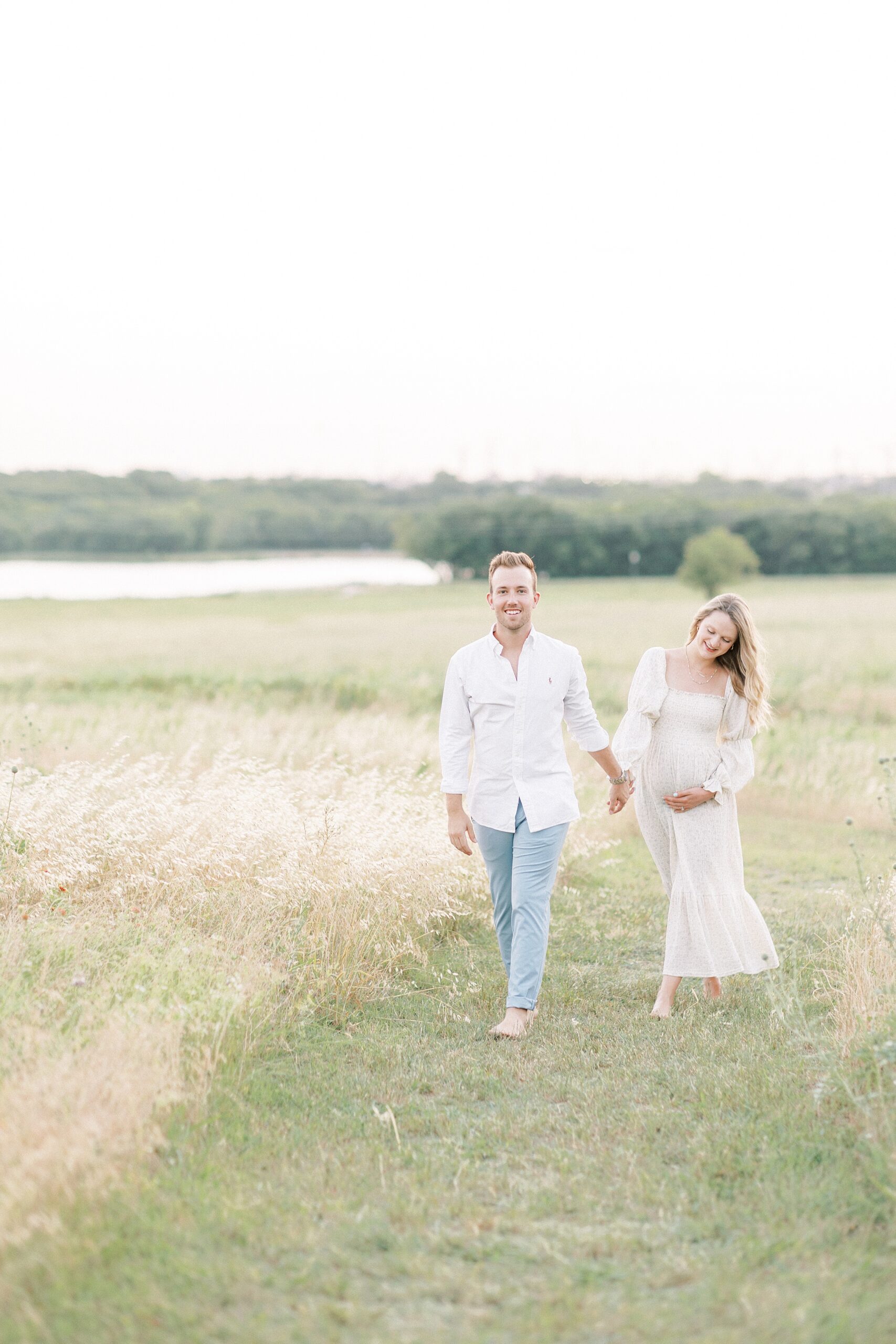 Happy expecting parents wearing white walk through a grassy field path holding hands before visiting Plano Birthplace